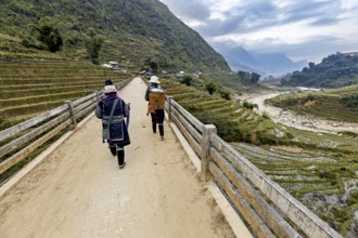 Two people walk on a bridge with a view of terraced rice paddies and mountains, local women farmers