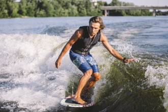 Young athletic man with long hair wakesurfing on waves of river in sunny summer weather. Ttheme