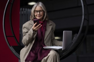 A senior businesswoman engages in work on her laptop outside, occasionally checking her smartphone.