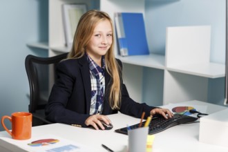 Little girl like business woman sitting at the desk with computer in blue office