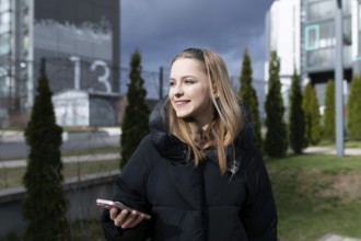 A young woman stands outdoors, smiling while using her smartphone. She is surrounded by modern