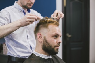 Barber Making Haircut Bearded Man In Barbershop. Professional stylist cutting client hair in salon.