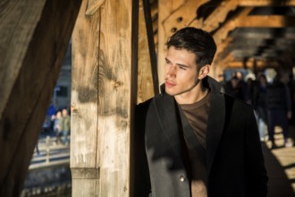 Young dark haired man standing on wooden medieval footbridge in Switzerland while looking away