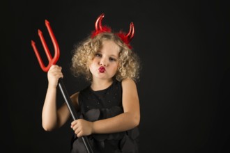 Isolated on black, adorable caucasian blonde little girl in black dress, with red horns on head