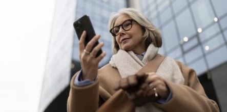 A mature woman wearing glasses and a warm coat is focused on her smartphone, standing on the