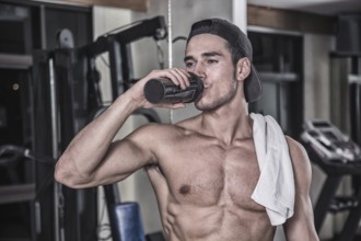 Attractive athletic shirtless young man drinking protein shake from blender in gym while looking at