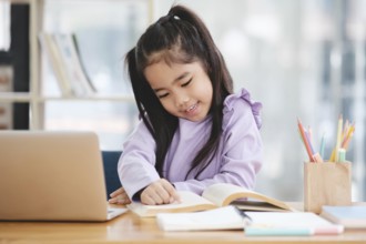A young girl is sitting at a desk with a laptop and a book. She is smiling and she is enjoying her