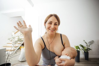 Portrait of cheerful mother in casual top nursing cute newborn while online meeting. Woman combines