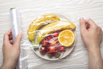 Male hands wrapping assorted different fruit and berries on white plate in plastic clear film top