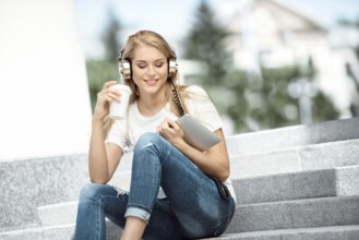 Happy young woman with vintage headphones, holding a takeaway coffee mug and a tablet PC, sitting