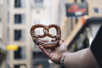 Woman hand holding a pretzel against cityscape. Focus on bread, blurred background