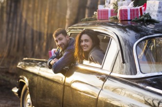 Loving man and woman sticking out of retro car windows with Christmas gifts on roof in park