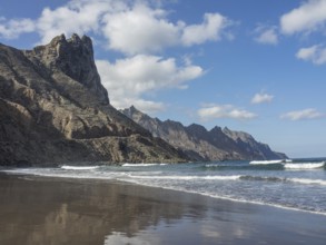 A peaceful beach with rocky cliffs and gently breaking waves under a blue sky and white clouds,