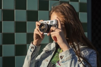 Cool girl model with retro film camera wearing a denim jacket, dark hair outdoors over city wall in