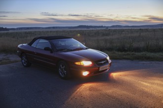 Convertible car parked on a road at sunset, casting an orange glow with fields around