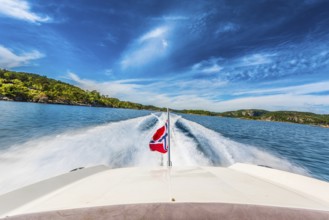 Speed boat swiftly crossing a sunny fjord in southern Norway sporting the Norwegian flag.