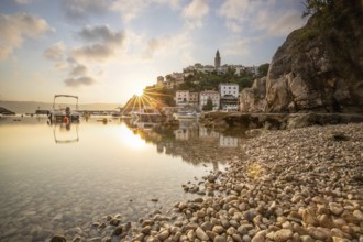 Harbour and beach with an old village on a hill. Boats are reflected in the water, the sun rises on