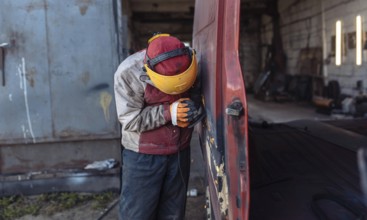 A worker sands a rusty van in an outdoor workshop setting