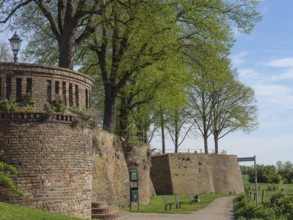 Defence defence tower and brick wall along a walkway, flanked by trees and park benches under a