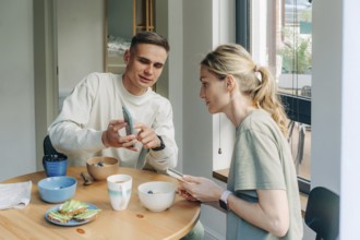 Ein Ehemann zeigt seiner Frau die Nachrichten auf seinem Handy, während er beim Frühstück am Tisch