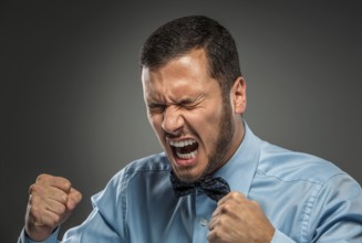 Portrait of angry upset young man in blue shirt and butterfly tie with fists up yelling isolated on
