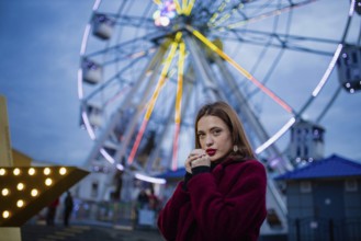 A thoughtful woman stands near a ferris wheel illuminated by colorful lights