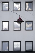 A hanging lamp in front of the façade of a residential building with windows reflecting the clouds