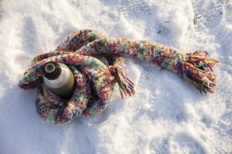 A long, colorful wool scarf lies on the snow. The scarf is wrapped around a thermos. The thermos is