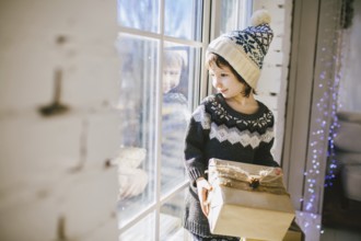 Child sitting at the window on a sunny Christmas day, making presents wrapped in boxes in paper.