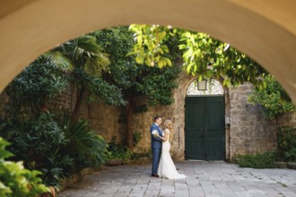 The bride and groom stand hugging in the coutryard in the old town of Perast, view through the arch