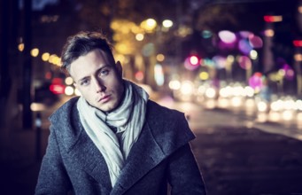 Handsome trendy young man, standing on a sidewalk in city setting at night wearing a fashionable