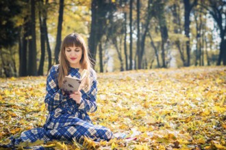Beautiful girl with long straight hair in a blue long dress reading a book in the autumn park