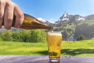 Hand of a man pouring beer from a bottle in a glass with a summer scenery in the background with