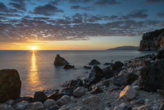 A colorful sunrise at Cala Gonone with black and white rocks and boulders and a sunburst