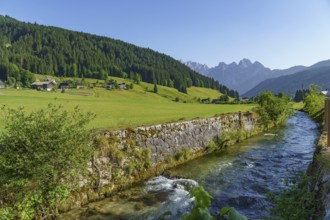 A clear river flows between wide green meadows and a mountain landscape under a bright blue sky,