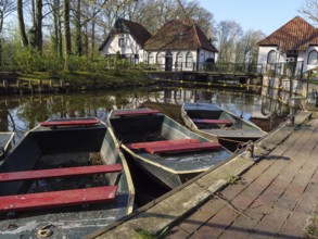 Boats in a pond in front of reflective houses with red roofs and white walls, surrounded by trees,