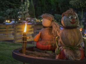 Two pumpkin sculptures sitting on a wooden wheel, illuminated by a burning torch in the evening