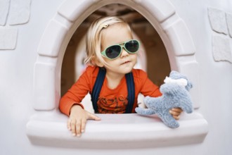 Little girl with a toy in her hand sits at the window of a toy house.