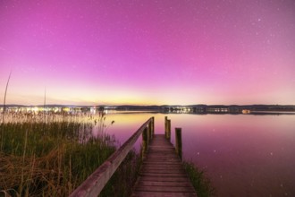 A wooden footbridge leads over a calm lake under a sky with aurora borealis, Reichenau, Lake