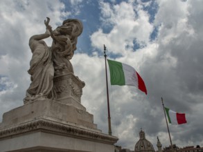 A statue and an Italian flag stand against a dramatic backdrop of clouds with ancient architecture