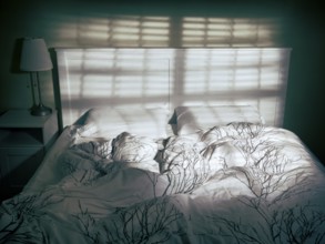 Unmade empty bed in dramatic dim light coming from a window through blinds
