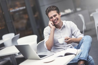 Handsome caucasian young man with phone working on laptop and smiling while enjoying coffee in cafe