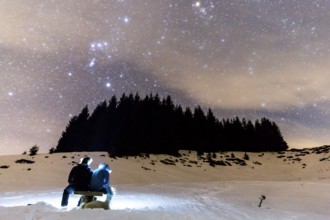 The Milky Way over the winter mountain landscape with pine trees. Two people sitting in the