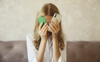 Desperate upset young woman holding mobile phone and bank card touching her head and covering face,