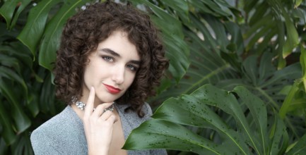 Woman with curly hair is standing in front of a green plant. She is wearing a blue dress and a
