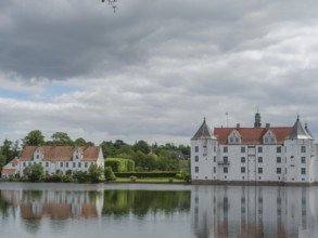 A historic castle with white facades and red roofs is reflected in a calm body of water under a