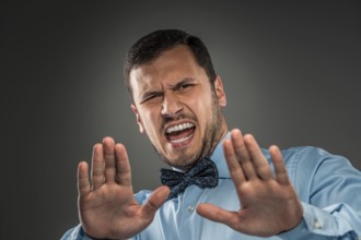Portrait of angry upset young man in blue shirt and butterfly tie with hands up yelling isolated on