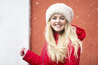 Portrait of a beautiful blonde girl in a warm hat and red jacket and falling snow