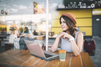 Beautiful young caucasian woman sitting on a terrace in a cafe in summer at a wooden table in a hat