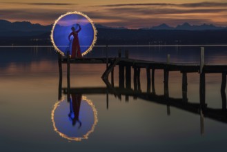 Woman in a red dress on a jetty at sunset on a lake, standing in front of a circle of light created